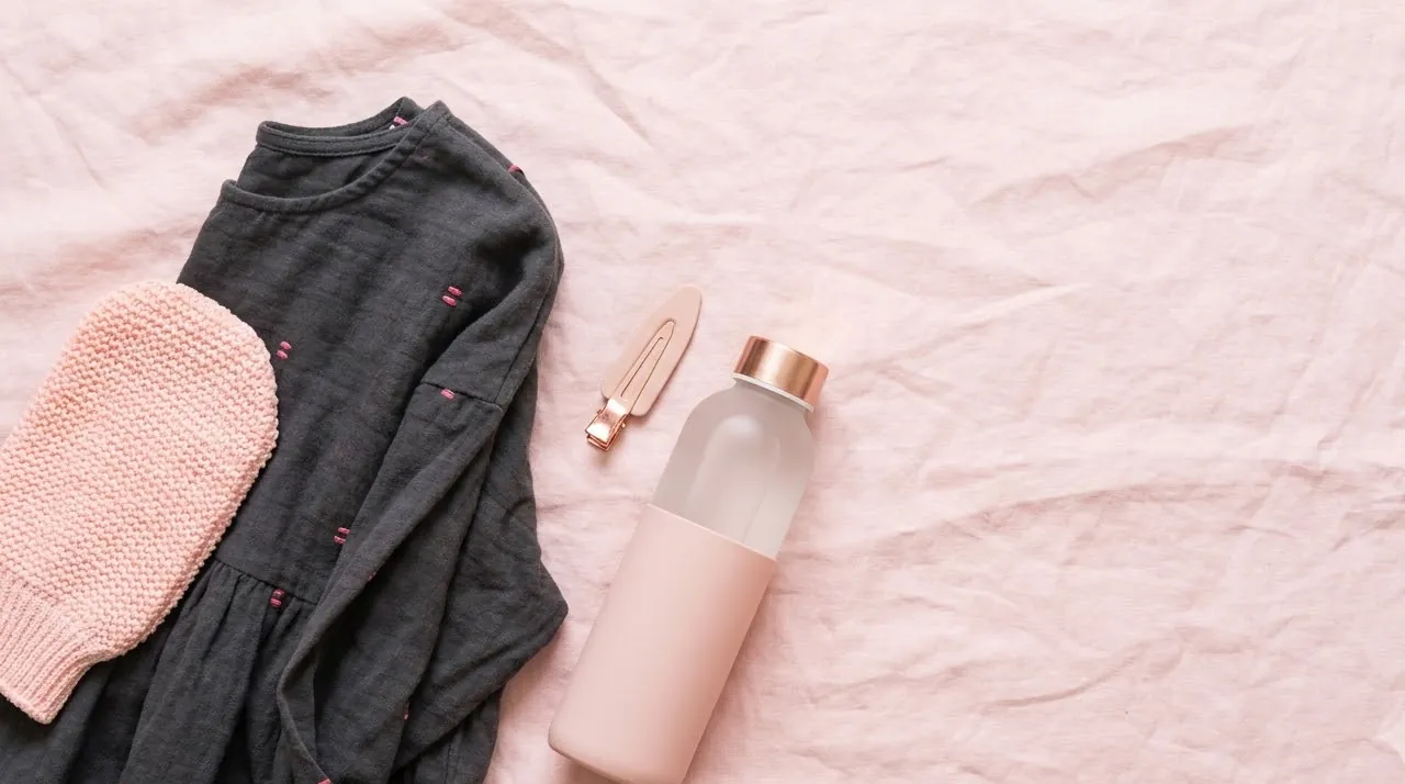 Flat-lay of exfoliating mitt, loose dark dress and water bottle — how to prepare for a spray tan